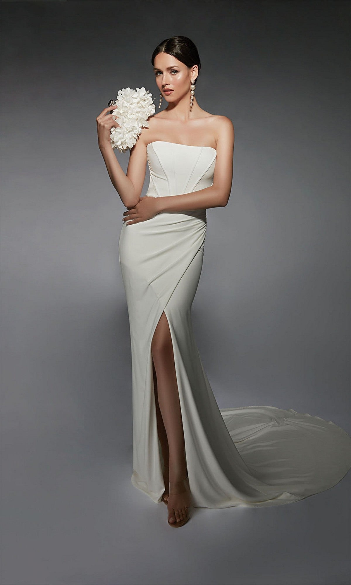 A woman barefoot poses with a white flower bouquet against a gray background, wearing the Long White Dress: Alyce 7102 featuring a strapless corset bodice and thigh-high slit.